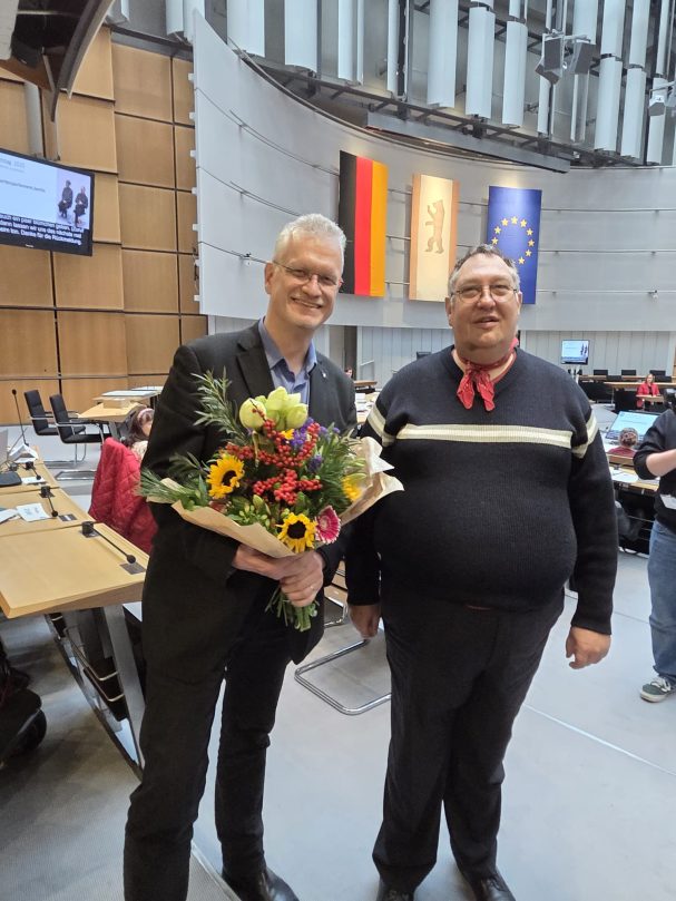 2 Männer lächeln in die Kamera. Einer hält einen Blumenstrauß. Im Hintergrund sind 3 Fahnen und der Plenarsaalt des AGH erkennbar.
