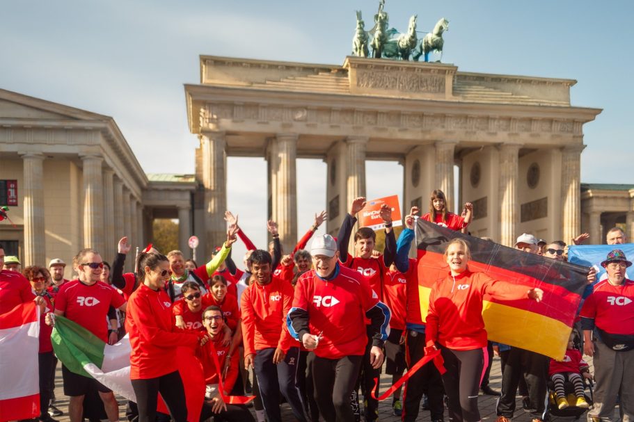 Gruppe von sportlich gekleideten Menschen mit einem roten Tshirt mit dem weißen Schriftzug RC halten Fahnen vor den Brandenburg Tor hoch.
