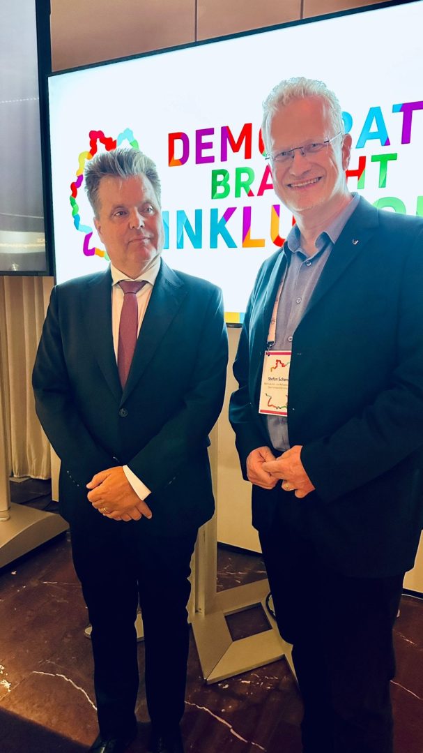 2 Herren in blicken in Richtung Kamera: Jürgen Dusel, und Stefan Schenck. Im Hintergrund ist der claim Demokratie braucht Inklusion 