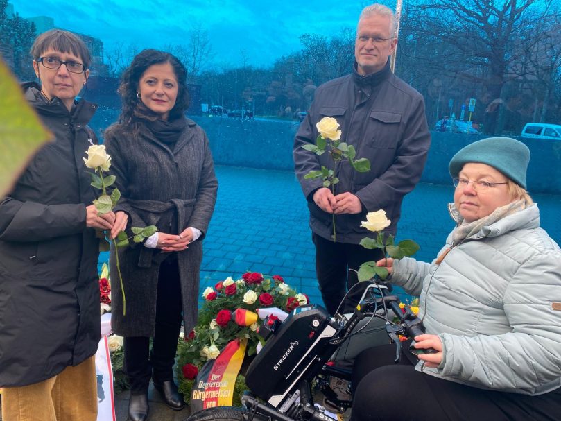 3 Frauen, davon eine im Rollstuhl sitzend, und Stefan Schenck stehen am Mahnmal der blauen Glaswand in der Tiergartenstraße 4 mit Blumen.